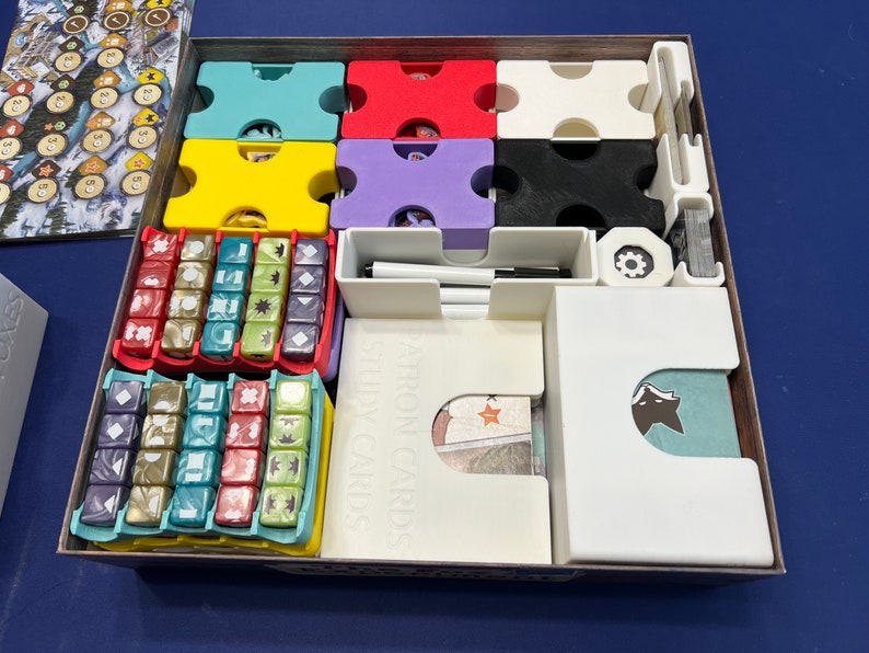 May include: A white box with dividers for organizing game components. The box contains a variety of colored dice, cards, and tokens. The box is labeled "Adventure Cards" and "Story Cards".