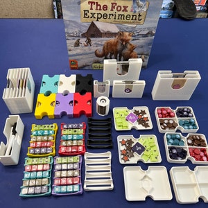 May include: A set of colorful plastic organizers for the board game "The Fox Experiment". The organizers are designed to hold the game's cards, tokens, and dice. The game box is also visible in the background.