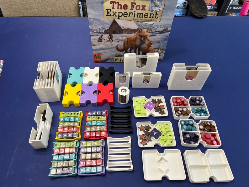 May include: A set of colorful plastic organizers for the board game "The Fox Experiment". The organizers are designed to hold the game's cards, tokens, and dice. The game box is also visible in the background.