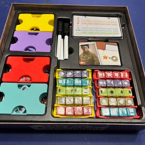 May include: The Fox Experiment board game box open, showing the game components: colorful dice, plastic tokens, a card with a woman in a military uniform, and a score sheet. The box lid has the game title "The Fox Experiment" printed on it.
