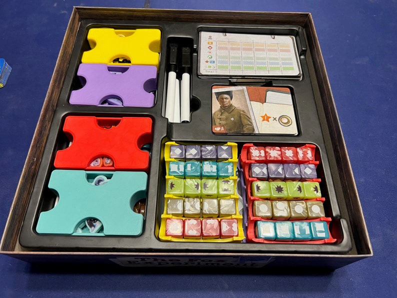 May include: The Fox Experiment board game box open, showing the game components: colorful dice, plastic tokens, a card with a woman in a military uniform, and a score sheet. The box lid has the game title "The Fox Experiment" printed on it.