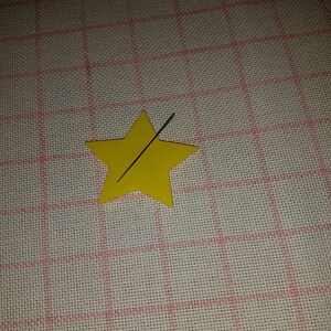 Peut inclure: Une étoile jaune vif à cinq branches en papier ou en carton, avec une fine aiguille à coudre métallique positionnée en diagonale au centre de l'étoile, reposant sur un fond à motif de grille blanche.