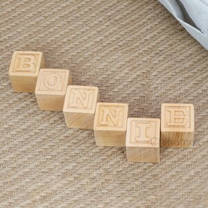 May include: Wooden blocks spelling out the name "BONNIE" in all caps. The blocks are arranged in a line on a brown woven surface.
