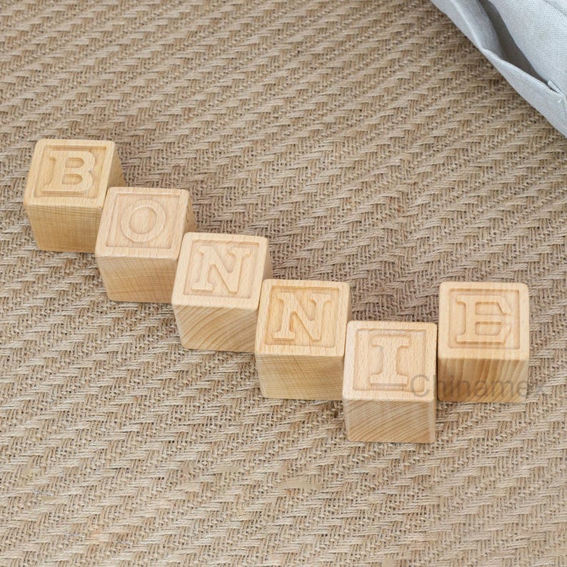 May include: Wooden blocks spelling out the name "BONNIE" in all caps. The blocks are arranged in a line on a brown woven surface.