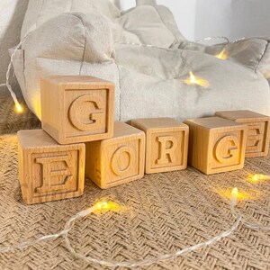 May include: Wooden blocks spelling out the name "GEORGE" in capital letters. The blocks are made of natural wood and have a slightly distressed finish. The blocks are arranged in a line on a woven surface.