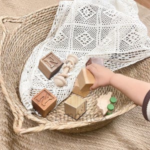 May include: Wooden blocks with the letter N on them, a wooden toy rattle, and a wooden toy dinosaur in a woven basket with a white crocheted blanket.