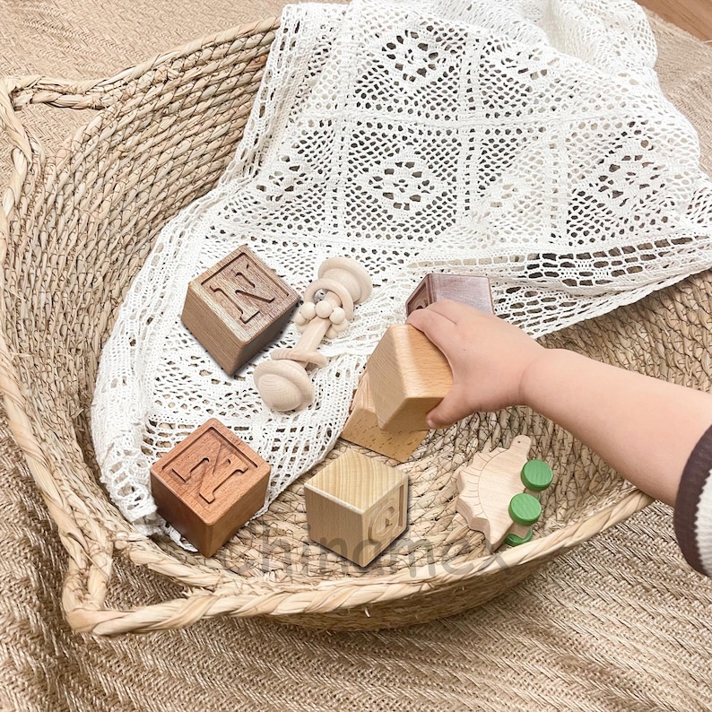 May include: Wooden blocks with the letter N on them, a wooden toy rattle, and a wooden toy dinosaur in a woven basket with a white crocheted blanket.
