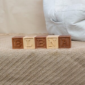May include: Wooden blocks spelling out the name "ELENA" in all capital letters. The blocks are arranged in a row on a brown fabric surface.