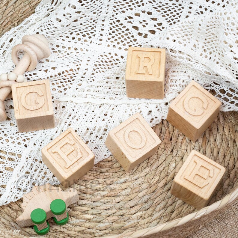 May include: Wooden blocks with the letters G, R, O, E, and H, arranged in a semi-circle on a woven basket surface. A small green toy is in the bottom left corner of the image.