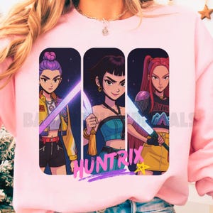 Może przedstawiać: Różowa bluza z nadrukiem przedstawiającym trzy kobiety w stylu anime trzymające świecące miecze. Pod postaciami znajduje się napis "HUNTRIX". Każda kobieta ma unikalną fryzurę i strój, z żywymi kolorami i futurystyczną estetyką.