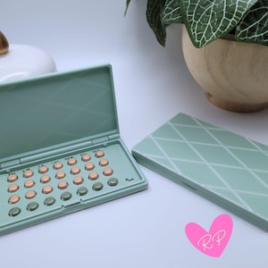 May include: A mint green pill case, open and closed, with a diamond pattern. The open case displays orange and green pills. A gold-topped white container and a plant in a wooden pot are in the background.