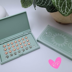 May include: A mint green pill case, open to reveal a set of pills, and a closed case with a sun and moon design. The open case shows pills in a grid, with days of the week labeled. The closed case has a decorative celestial design.
