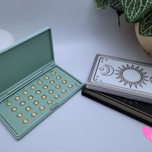 May include: Two pill cases, one green and one white with a gold sun and moon design. The green case is open and contains a full pack of pills.