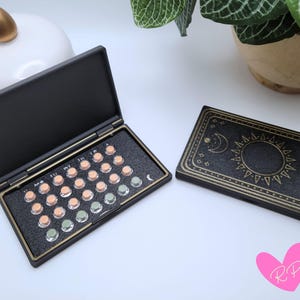 May include: A black pill case with a gold trim, open to reveal a series of pills. Next to it is a closed black case with a gold sun and moon design. The cases are on a white surface.