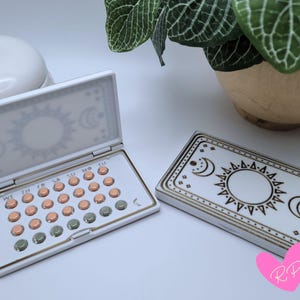 May include: A white pill case with a celestial design, open to reveal a 28-day cycle of pills. The case is decorated with a sun and moon motif. A closed matching case is also present.