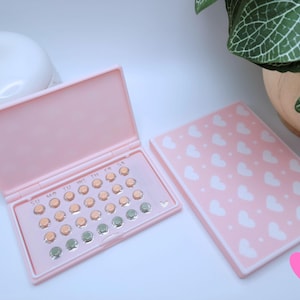 May include: A pink pill case, open to reveal a grid of pills in shades of orange and green. The case is next to its matching pink cover, decorated with white hearts. A white container with a gold egg-shaped top is in the background.