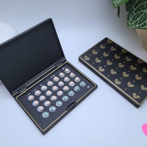 May include: Black rectangular case with a gold trim, open to reveal a grid of small, round pills in shades of pink and blue. A matching closed case with gold heart pattern is next to it. The cases are on a white surface.