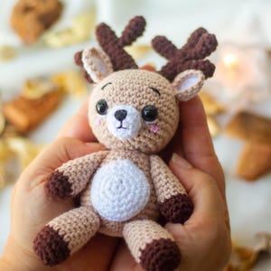 May include: A crocheted reindeer toy with brown antlers, brown hooves, and a white belly. The toy is being held in a person's hands.