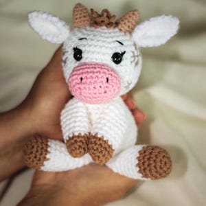 May include: A white crocheted cow plush toy with brown accents. The cow has a pink nose and black eyes. The toy is being held in a person's hands.
