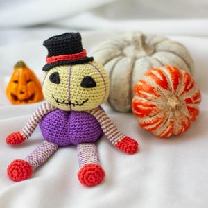 May include: A crocheted pumpkin doll wearing a black top hat with a red band. The doll has a purple body, yellow face, and black eyes. The doll is sitting on a white surface with two pumpkins.