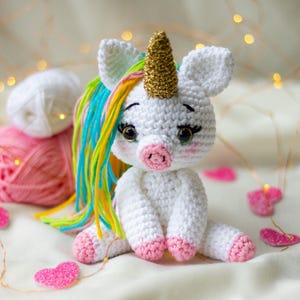 Può includere: Un unicorno bianco fatto a mano all'uncinetto con un corno dorato, criniera arcobaleno e zoccoli rosa. L'unicorno è seduto su una superficie bianca con decorazioni a forma di cuore rosa e gomitoli di lana sullo sfondo.