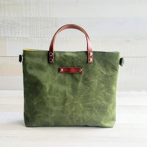 May include: A green canvas tote bag with brown leather handles and a brown leather label. The bag has a yellow lining.