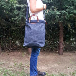 May include: A dark blue canvas tote bag with brown leather handles and a long adjustable strap. The bag is worn over the shoulder of a person wearing blue jeans and a white t-shirt.
