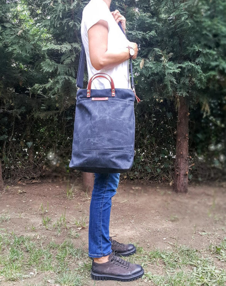 May include: A dark blue canvas tote bag with brown leather handles and a long adjustable strap. The bag is worn over the shoulder of a person wearing blue jeans and a white t-shirt.