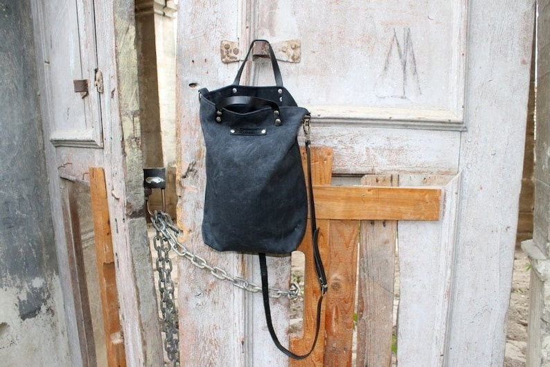 Op de afbeelding: Een zwarte canvas tote bag met leren banden en een lange schouderband. De tas hangt aan een houten deur met een ketting en een slot.
