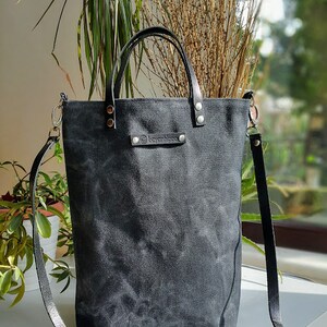 Op de afbeelding: Een zwarte canvas tote bag met een leren band en een leren handvat. De tas heeft een klein leren label met het woord "Kyanibingo" erop.