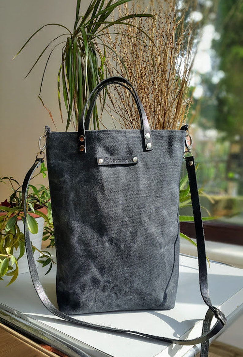 Op de afbeelding: Een zwarte canvas tote bag met een leren band en een leren handvat. De tas heeft een klein leren label met het woord "Kyanibingo" erop.