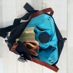 May include: A black canvas tote bag with brown leather handles and a blue lining. The bag is open and filled with books, a phone, and a water bottle.