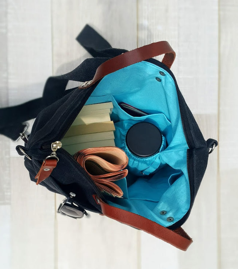 May include: A black canvas tote bag with brown leather handles and a blue lining. The bag is open and filled with books, a phone, and a water bottle.