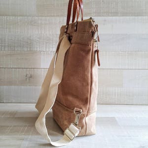 May include: A brown canvas tote bag with a white strap and brown leather handles. The bag has a zipper closure and a small pocket on the front.