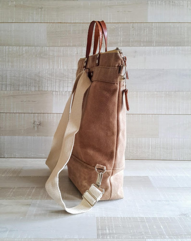 May include: A brown canvas tote bag with a white strap and brown leather handles. The bag has a zipper closure and a small pocket on the front.