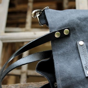 Op de afbeelding: Een donkergrijze canvas tote bag met zwarte leren banden en zilveren hardware. De tas heeft een label met de tekst "bagahjiba" in witte letters.