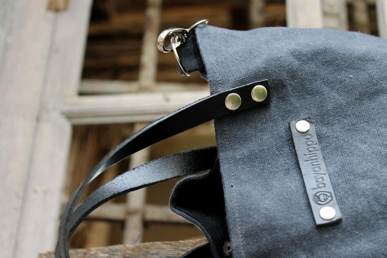 Op de afbeelding: Een donkergrijze canvas tote bag met zwarte leren banden en zilveren hardware. De tas heeft een label met de tekst "bagahjiba" in witte letters.
