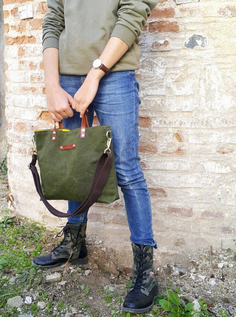May include: A green canvas tote bag with brown leather handles and a brown adjustable shoulder strap. The bag has a yellow lining and a brown leather patch on the front.