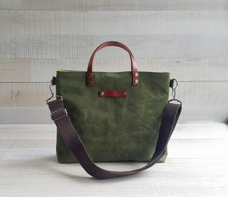 May include: A green canvas tote bag with brown leather handles and a brown adjustable shoulder strap. The bag has a zipper closure and a small pocket on the front.
