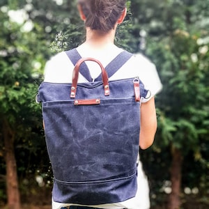 May include: A dark blue canvas backpack with brown leather straps and handles. The backpack has a zipper closure and a flap with a metal buckle.