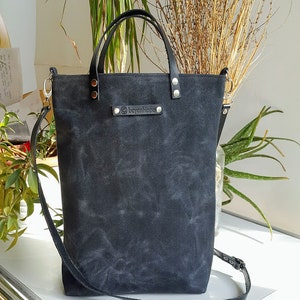 Op de afbeelding: Zwarte canvas tote bag met leren handvatten en een zwarte leren schouderband. De tas heeft een klein leren label met de tekst "boyanipsoo" erop.