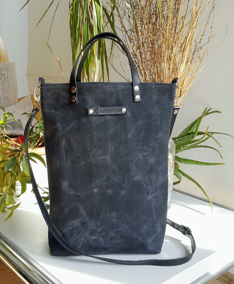 Op de afbeelding: Zwarte canvas tote bag met leren handvatten en een zwarte leren schouderband. De tas heeft een klein leren label met de tekst "boyanipsoo" erop.