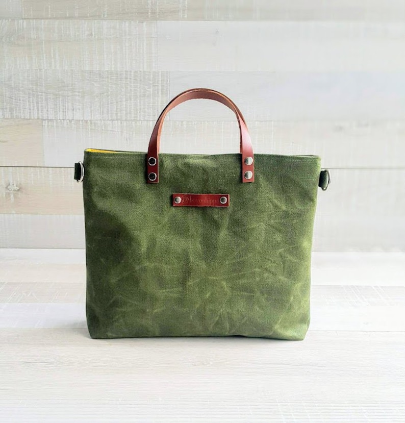 May include: A green canvas tote bag with brown leather handles and a brown leather label. The bag has a yellow lining.