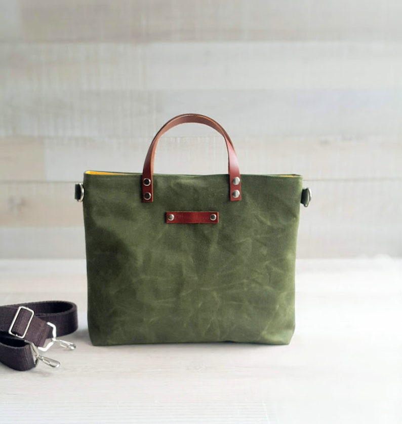 May include: A green canvas tote bag with brown leather handles and a brown leather strap. The bag has a yellow lining and silver hardware.