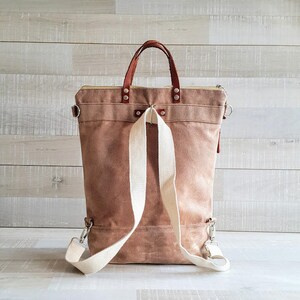 May include: A brown canvas backpack with a leather handle and white straps. The backpack has a zipper closure and a small pocket on the front.