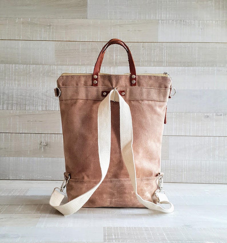 May include: A brown canvas backpack with a leather handle and white straps. The backpack has a zipper closure and a small pocket on the front.