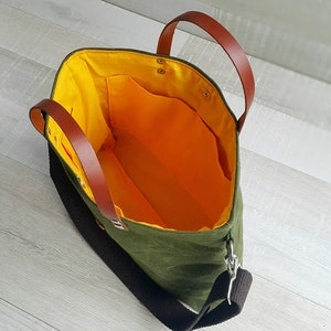 May include: A green canvas tote bag with brown leather handles and a black adjustable shoulder strap. The bag has a yellow lining and a small pocket inside.