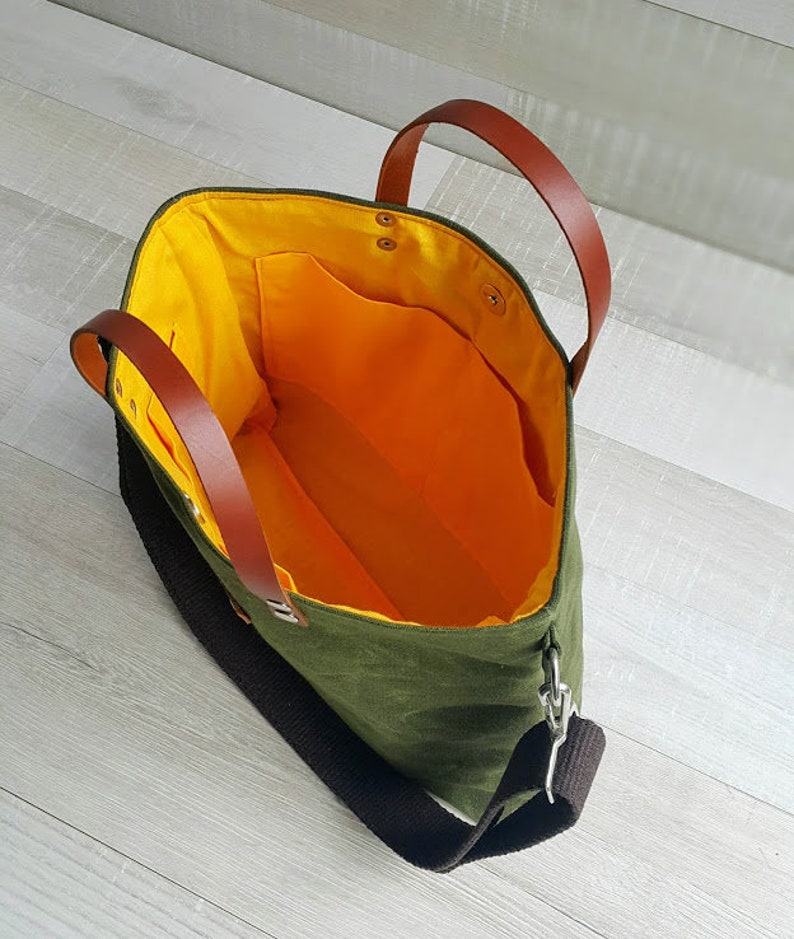 May include: A green canvas tote bag with brown leather handles and a black adjustable shoulder strap. The bag has a yellow lining and a small pocket inside.