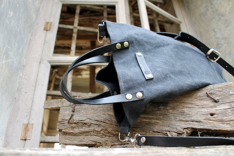 Op de afbeelding: Een zwarte canvas tote bag met leren banden en een zilveren gesp. De tas heeft een klein leren patch met het woord "REBEL" erop.