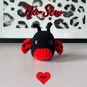 Op de afbeelding: Een zwart en rood gehaakt lieveheersbeestje speelgoed met witte ogen en antennes. Het lieveheersbeestje heeft rode vleugels en een rood onderlichaam. De tekst "No-Sew" is in rood script boven het lieveheersbeestje geschreven. Een rood hart met "PDF" staat onderaan.